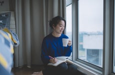Asian student sitting by the window of her apartment looking out over the city with a pen and notepad in one hand and a cup of coffee in the other.