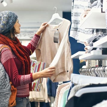 Closeup of smiling blond woman choosing clothes at department store in local supermarket. She’s holding a beige blouse and looking at it. The woman is wearing gray cap, red sweater and scarf. Side view.