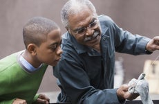 Older black grandpa is changing the oil on his car and showing his grandson what he's doing.