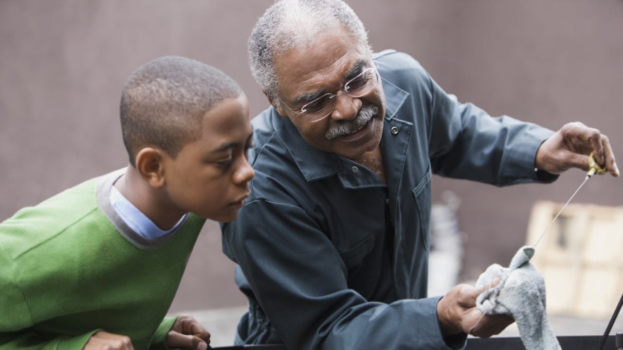 Older black grandpa is changing the oil on his car and showing his grandson what he's doing.