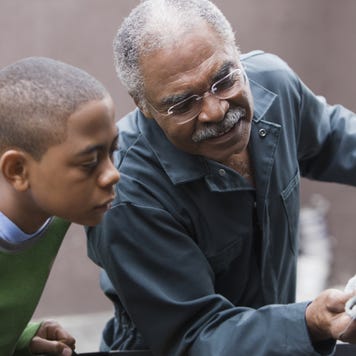 Older black grandpa is changing the oil on his car and showing his grandson what he's doing.