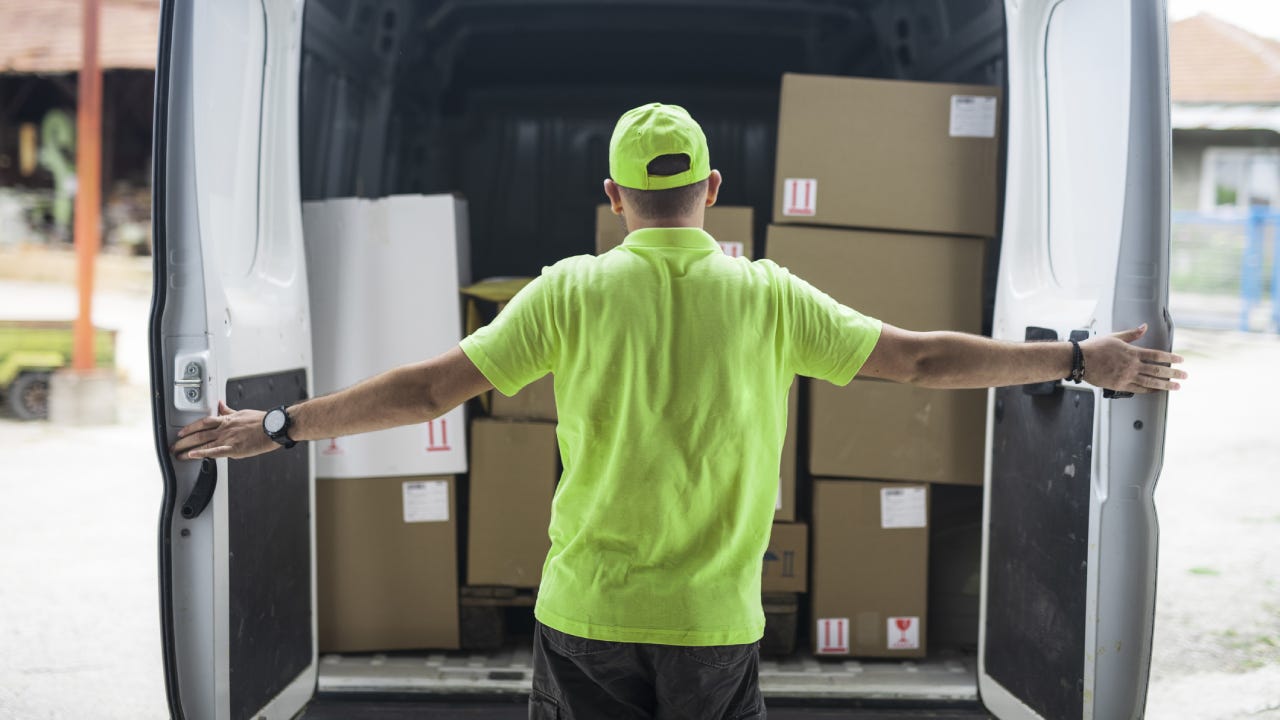 Working man in neon hat and shirt opens the rear doors to his delivery van revealing stacks of boxes for delivery.