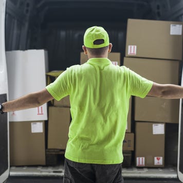 Working man in neon hat and shirt opens the rear doors to his delivery van revealing stacks of boxes for delivery.
