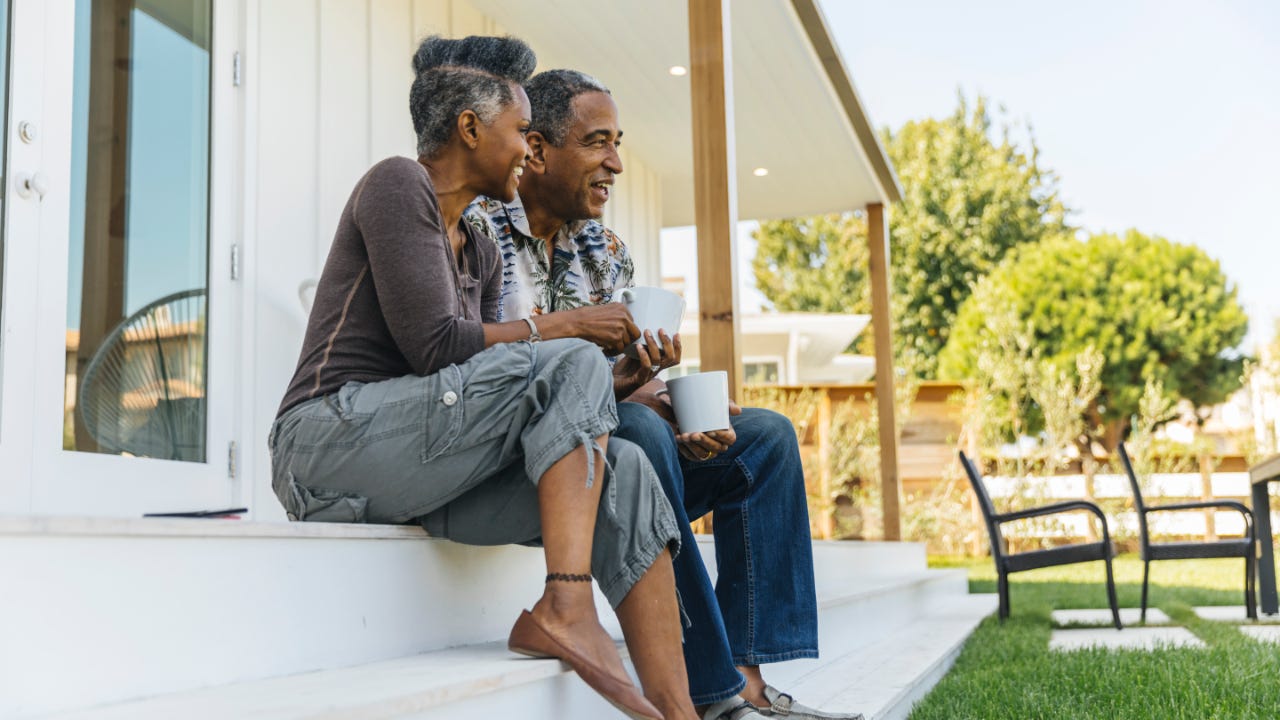 An older Black couple sits on their porch and drinks coffee
