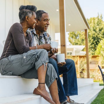 An older Black couple sits on their porch and drinks coffee