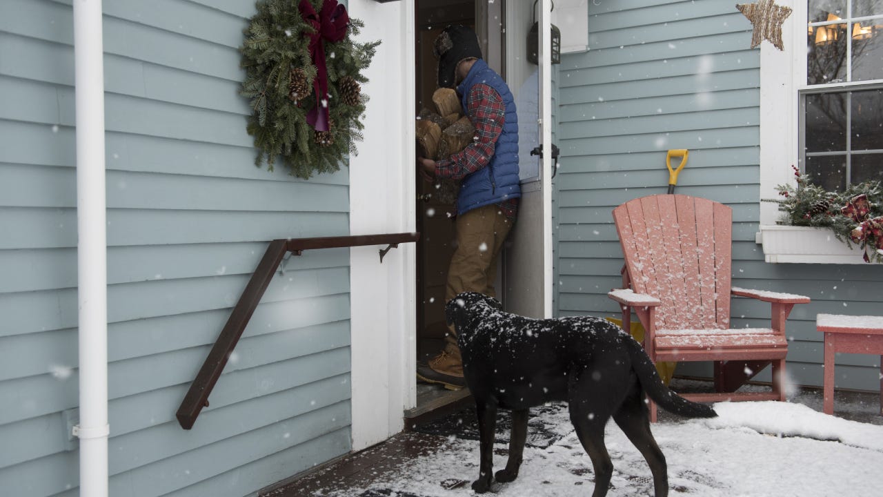 A man holding an armful of chopped wood enters his home while his black dog stands outside on the deck wanting to play. It is snowing.