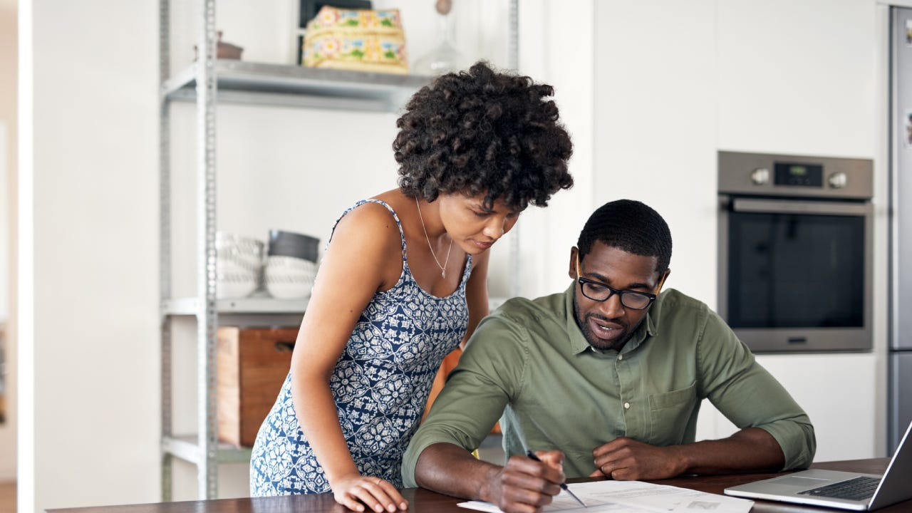 Black couple together in the kitchen at the table. The husband is sitting down while the wife is standing over him and looking at the papers on the table with him.