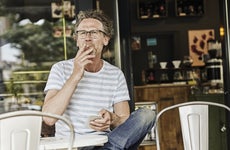 An older man with glasses sitting at a coffee shop outside and smoking a cigarette while he holds his smartphone.
