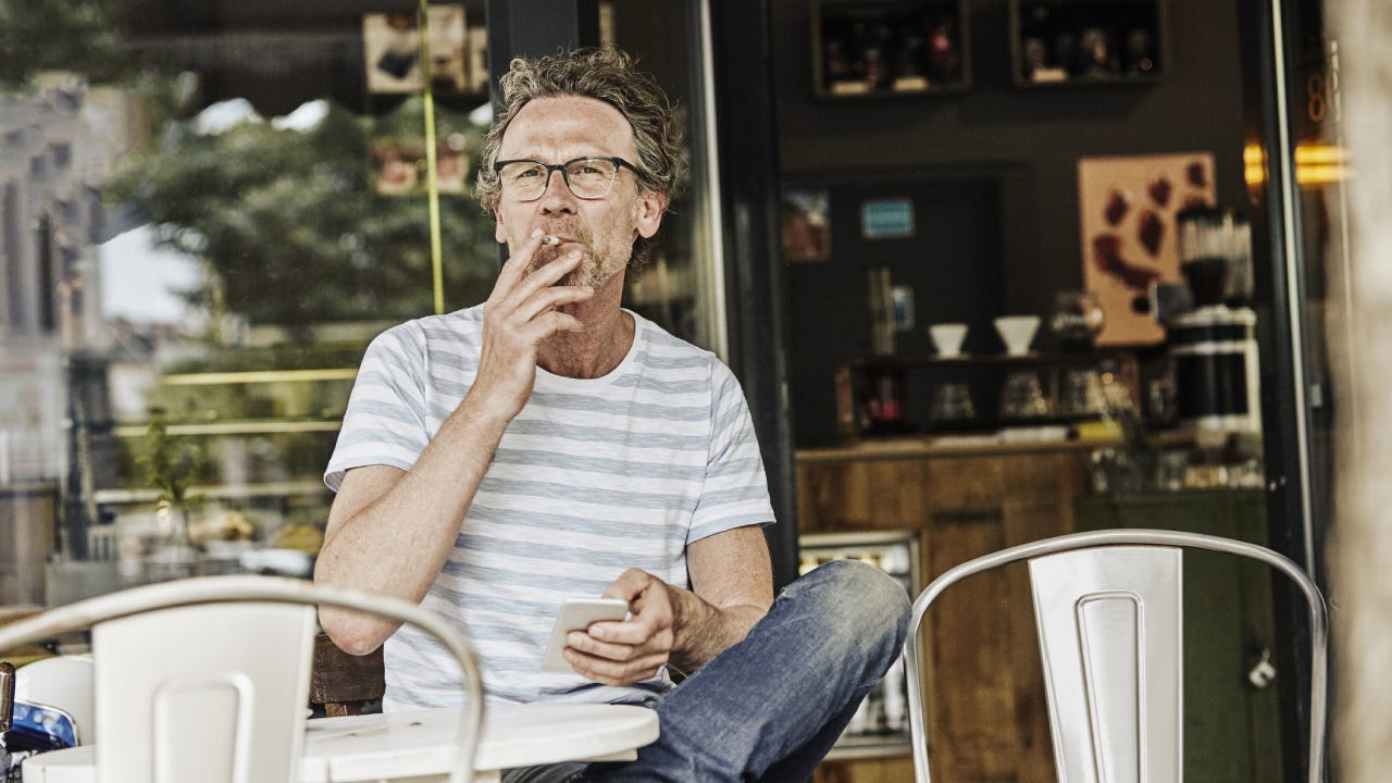 An older man with glasses sitting at a coffee shop outside and smoking a cigarette while he holds his smartphone.