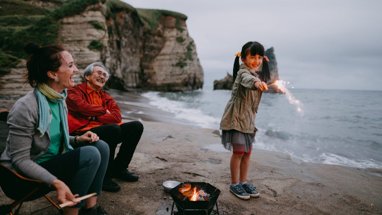A mixed-race Asian girl stands with her grandparents (one white woman and one Asian man) on the beach playing with a sparkler. It is dusk.
