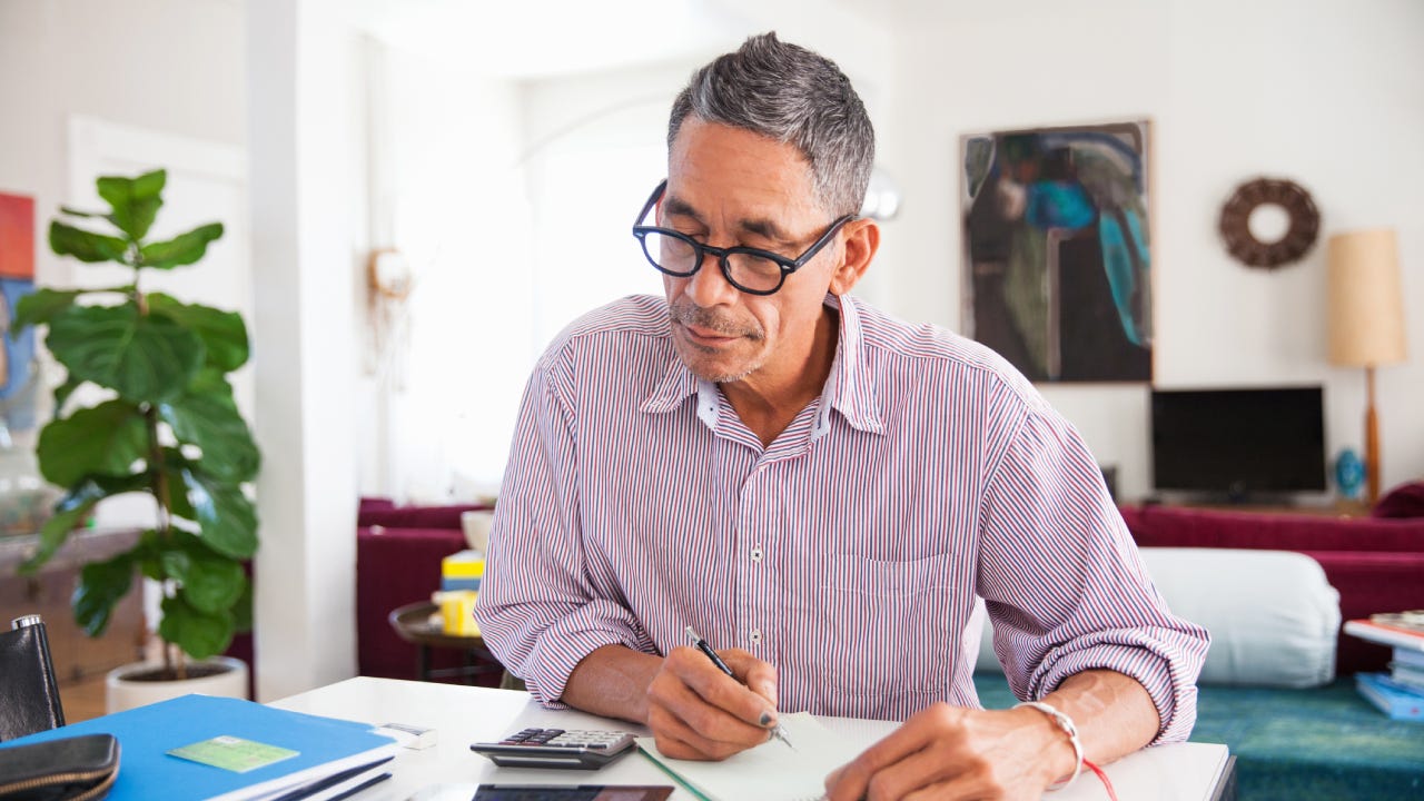 An older man sits down at his table with some papers and a calculator to review his finances after a home insurance claim.