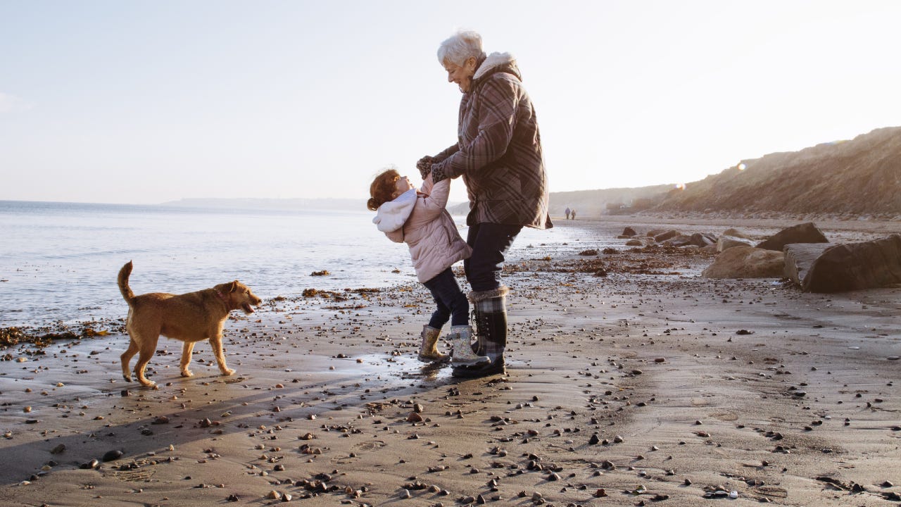 Grandmother plays with her granddaughter on the beach while a dog runs around the shore.