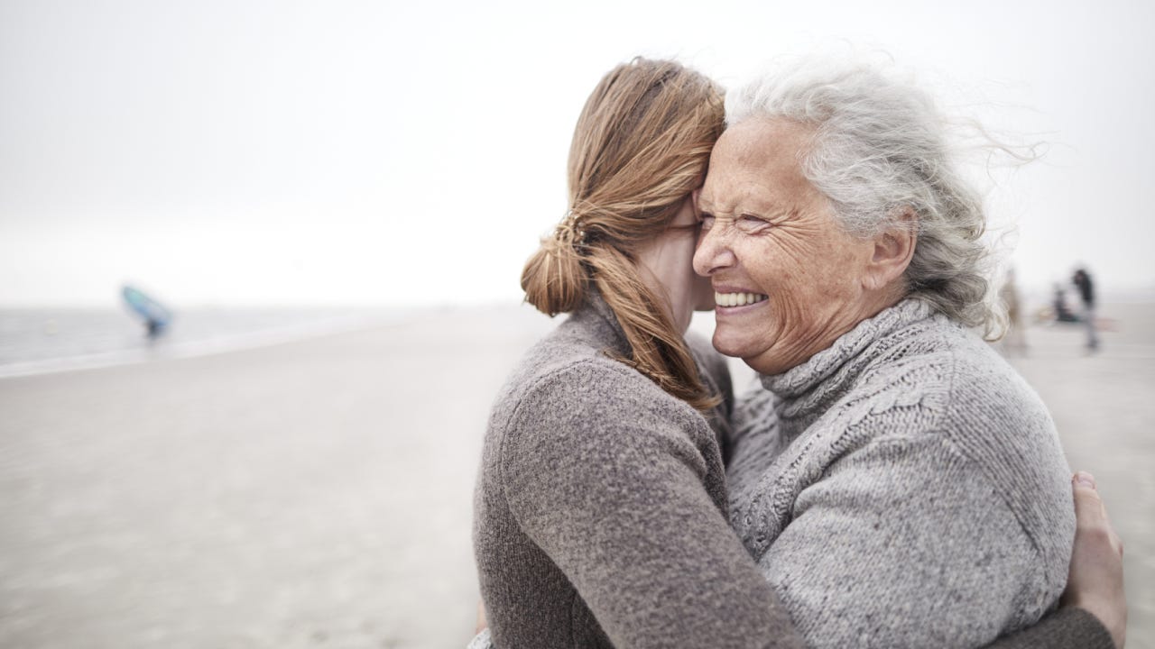 An old mother hugs her adult daughter on a beach.