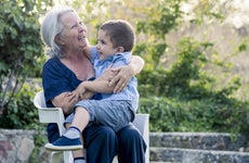 A grandma sits on a chair with her grandson in her lap as they laugh together outside.