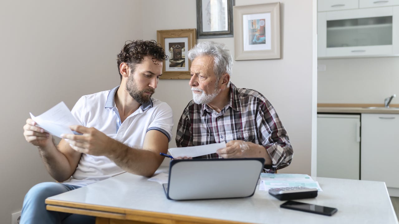 An adult son goes over some policy forms with his father while sitting in front of a laptop.