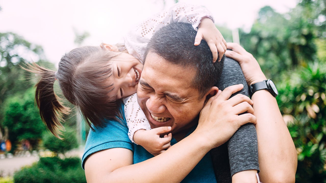 A middle-aged Asian man has his little daughter on his shoulders and they are playing and laughing.