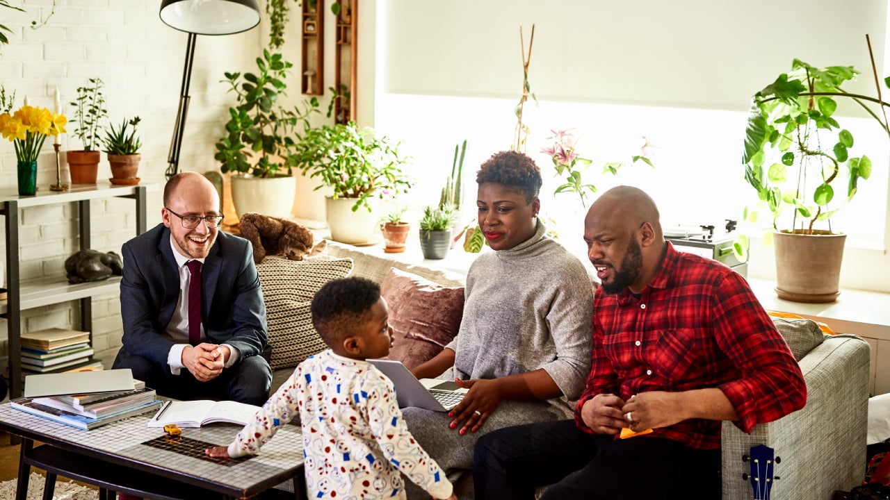 A black couple is sitting together on a couch in the middle of talking to a financial advisor when their son shows up in his pajamas to ask a quesiton.