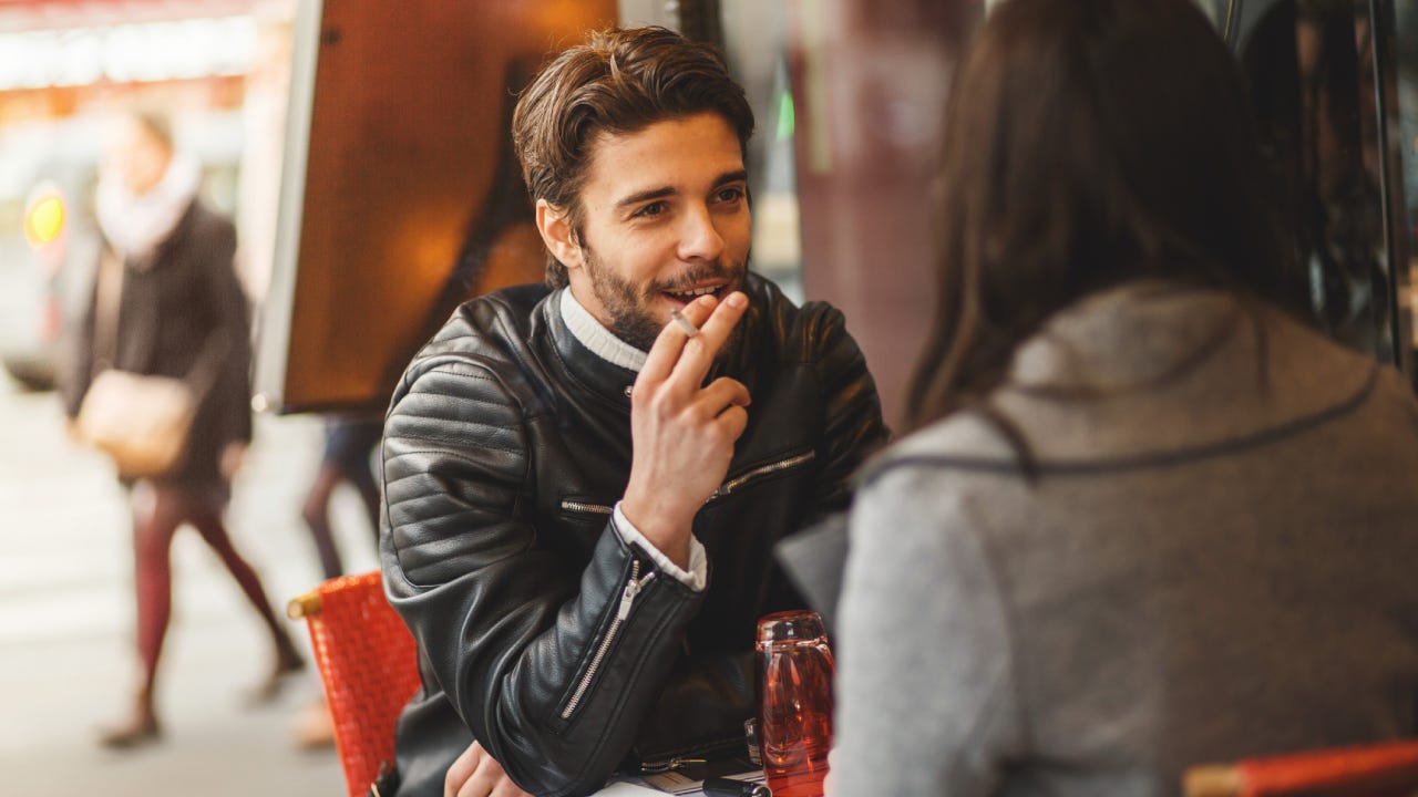 A man and a woman are out at a cafe. The man is holding a cigarette between his fingers.