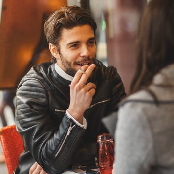 A man and a woman are out at a cafe. The man is holding a cigarette between his fingers.