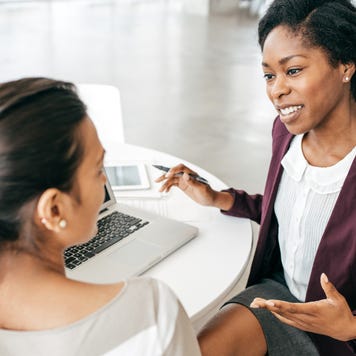 A black lady who is a financial advisor is sitting down on a white table across from a Caucasian woman client. They are talking about something and a laptop is open in the table.