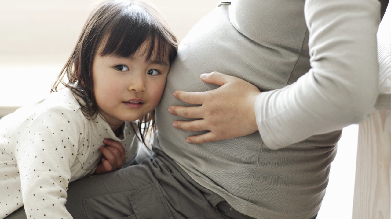 A little Asian girl puts her ear to her pregnant mother's belly to hear the baby inside.