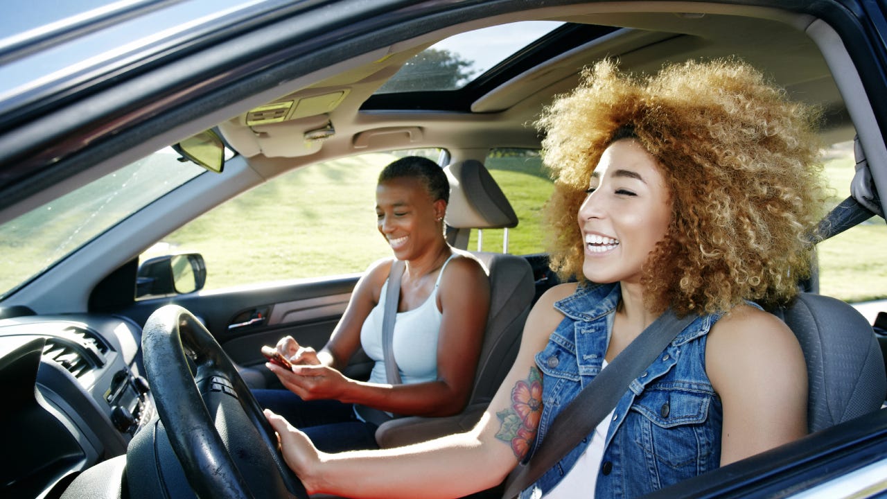 Black daughter drives while her mother is in the passenger side on the phone. They are both laughing and smiling.