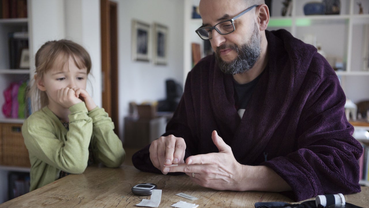 A dad checks his blood with a diabetic testing kit while his daughter watches, confused.