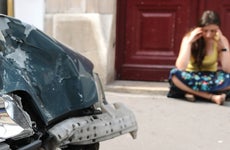 A woman sits on the sidewalk on her smartphone next to her car that has been hit.