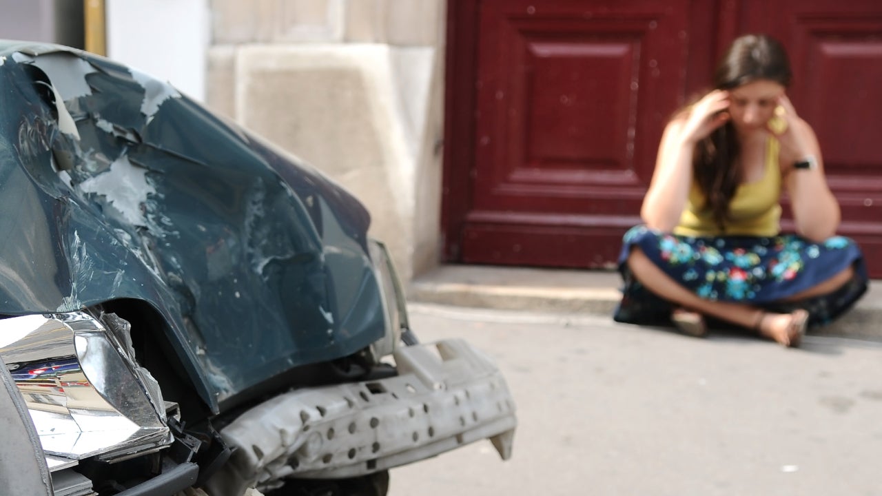 A woman sits on the sidewalk on her smartphone next to her car that has been hit.