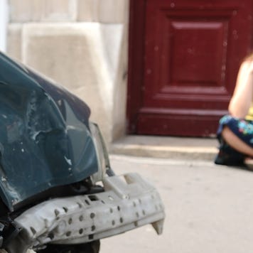 A woman sits on the sidewalk on her smartphone next to her car that has been hit.