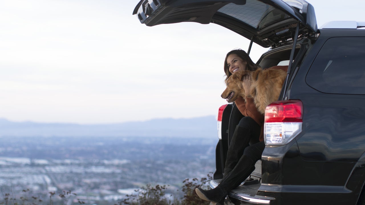 A woman sits in the back of her SUV with the back open and her dog is sitting with her.
