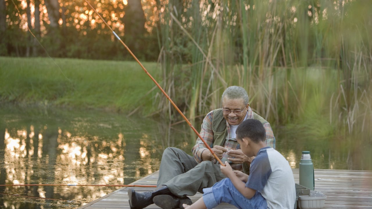 Black senior and his grandson are sitting together on a dock with fishing rods laughing together.