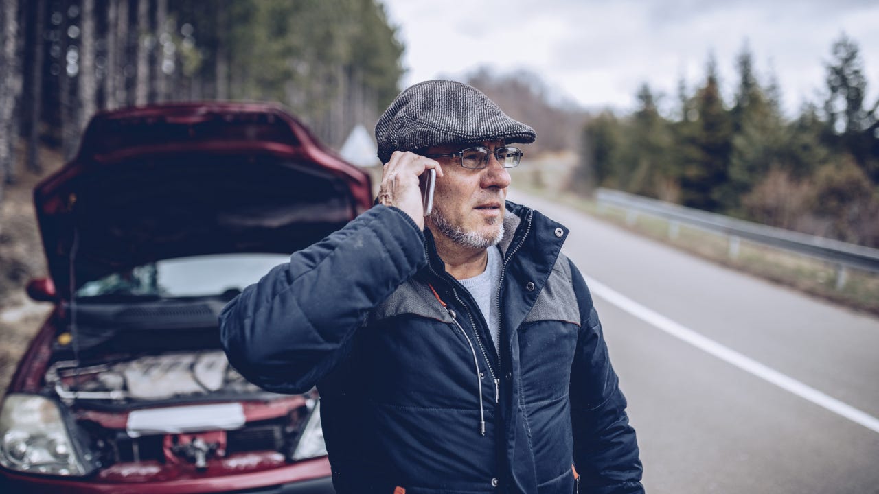 An older man standing on the side of the road on his phone beside his car which has the hood up.