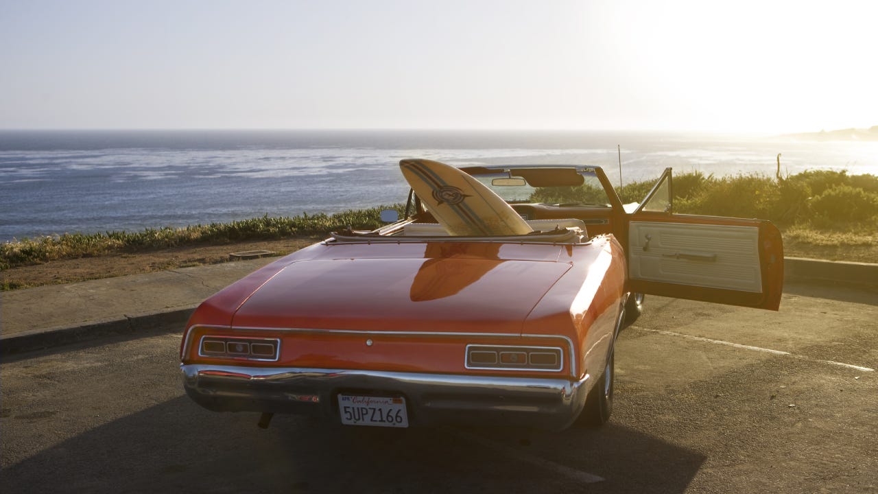 A red convertible on the coast with a surf board sticking out of it.