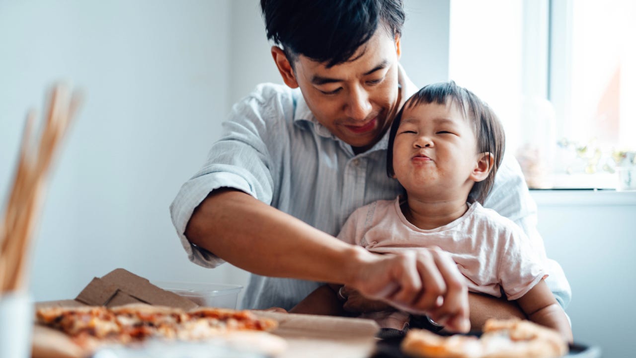 A father sits with his young child feeding him a piece of pizza.