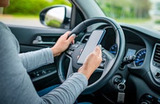 Shot of a driver's hands as they sit in the driver's seat with one hand on the steering wheel and one hand holding up a smartphone.
