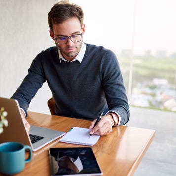 Man sitting at laptop taking down finance notes by hand