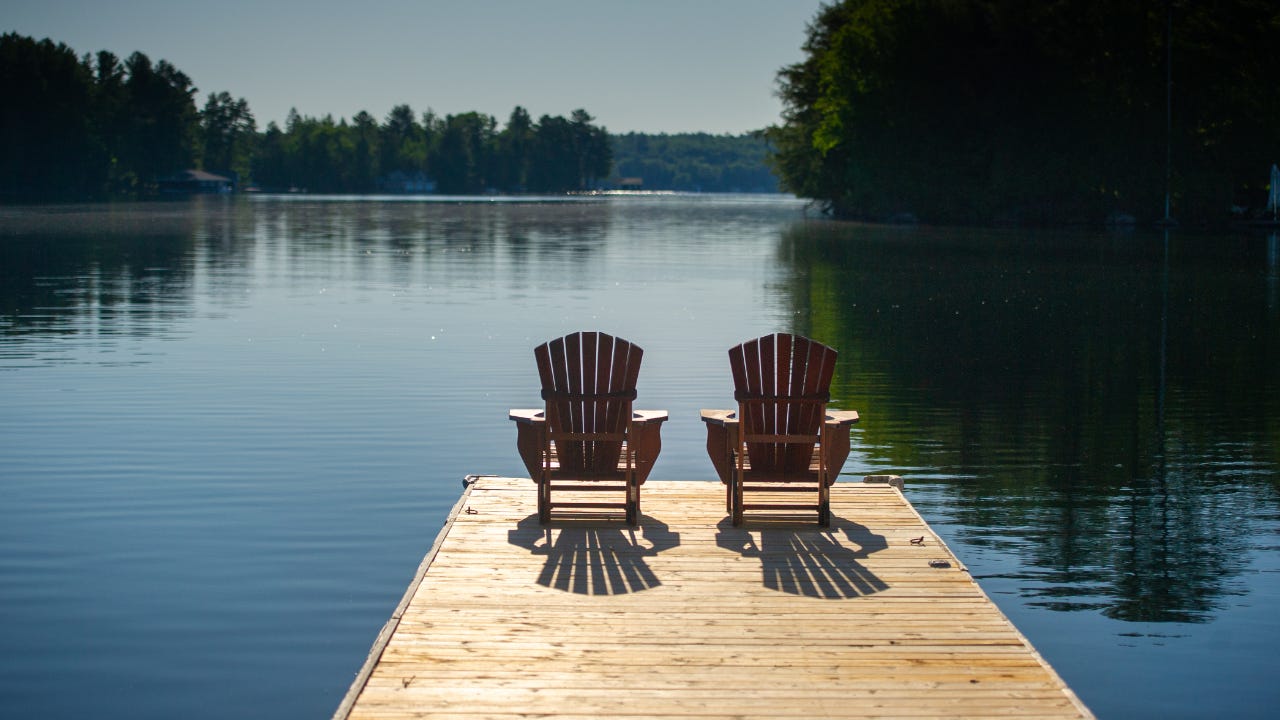 Two Adirondack chairs sitting on a wooden pier facing the calm water of a lake in Muskoka, Ontario Canada. A cottage nestled between trees is visible in background