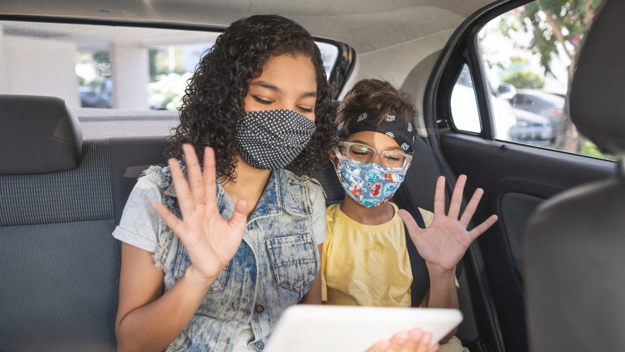 Two sisters are wearing facemasks and waving to someone on their tablet while sitting in a car