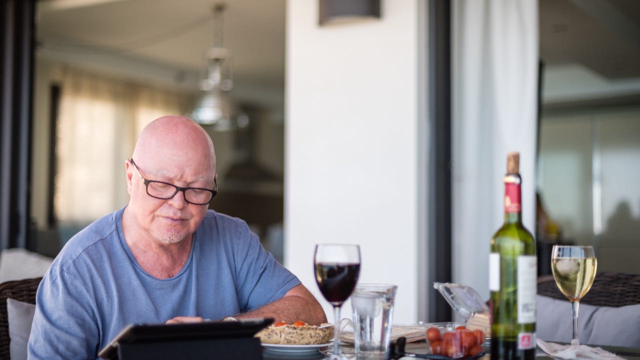 An older man researches online while having a glass of wine.