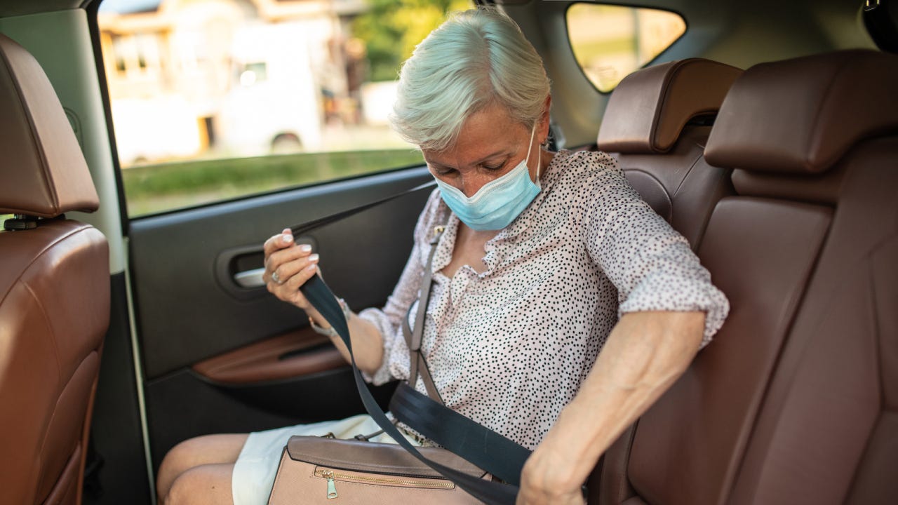 Senior woman wearing protective face mask, fastening seat belt before ride in ride sharing car