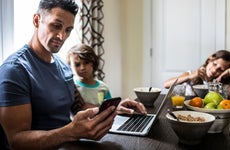 father checking phone and computer at breakfast table