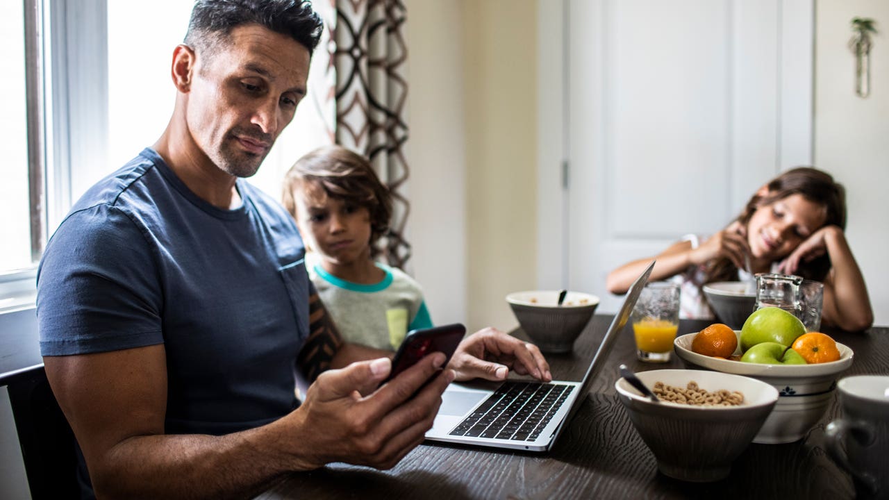 father checking phone and computer at breakfast table