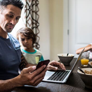 father checking phone and computer at breakfast table
