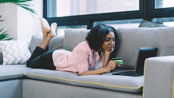 Black woman laying on couch using laptop and holding credit card