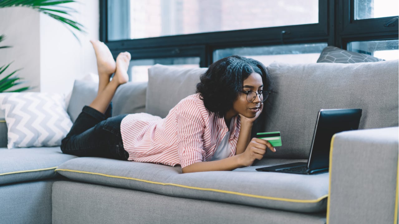 Black woman laying on couch using laptop and holding credit card