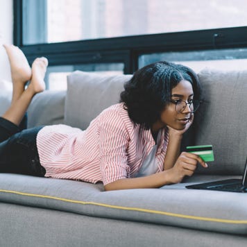 Black woman laying on couch using laptop and holding credit card