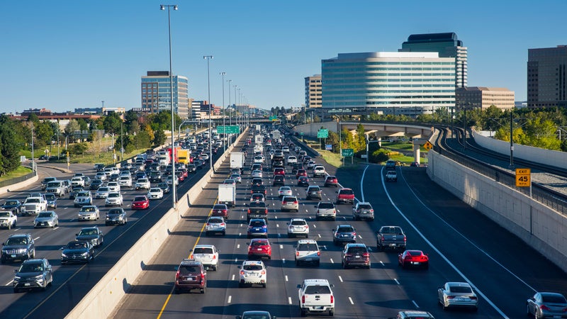 A busy freeway full of cars of various shapes and colors on a bright, sunny day with buildings in the background.
