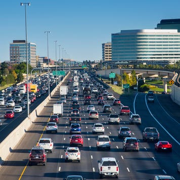 A busy freeway full of cars of various shapes and colors on a bright, sunny day with buildings in the background.