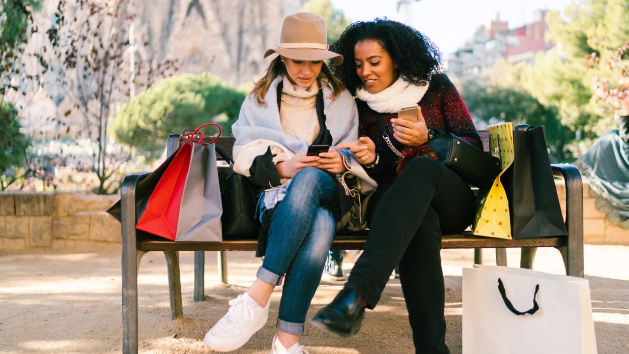 Two young women sit on bench with shopping bags looking at one cell phone
